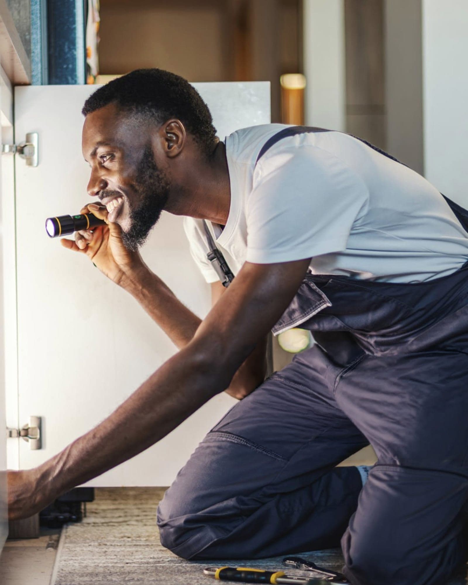 A leak detection specialist kneels on the kitchen floor of a Butte County home, shining a flashlight under a cabinet to inspect for hidden leaks beneath the sink, with a screwdriver and inspection tools laid out beside him.