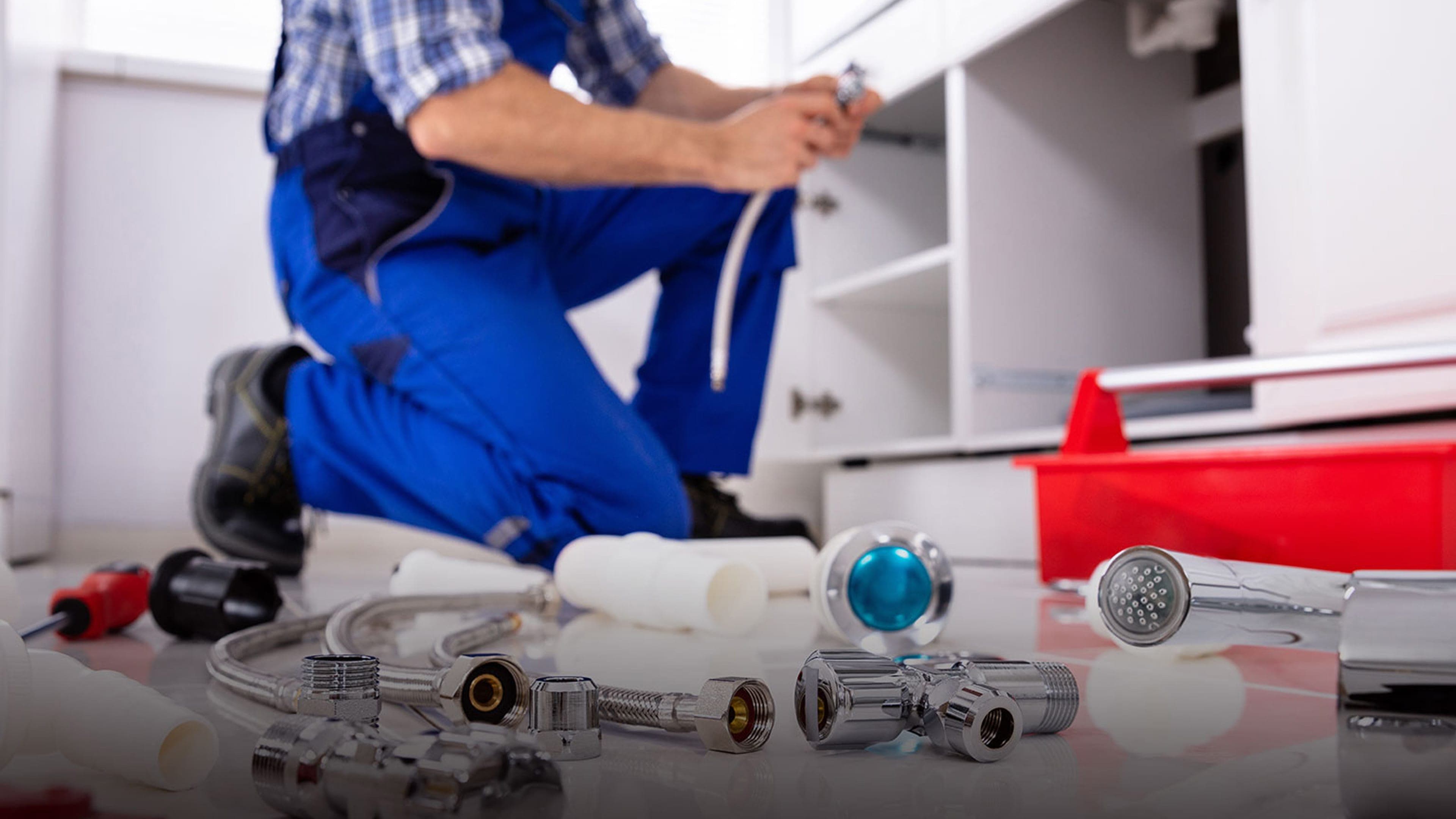 A leak detection specialist kneels under a residential kitchen sink cabinet in Sierra County, with chrome plumbing fittings, faucet components, valves, and a red tool tray laid out on the floor in the foreground, a service call mid-diagnosis.