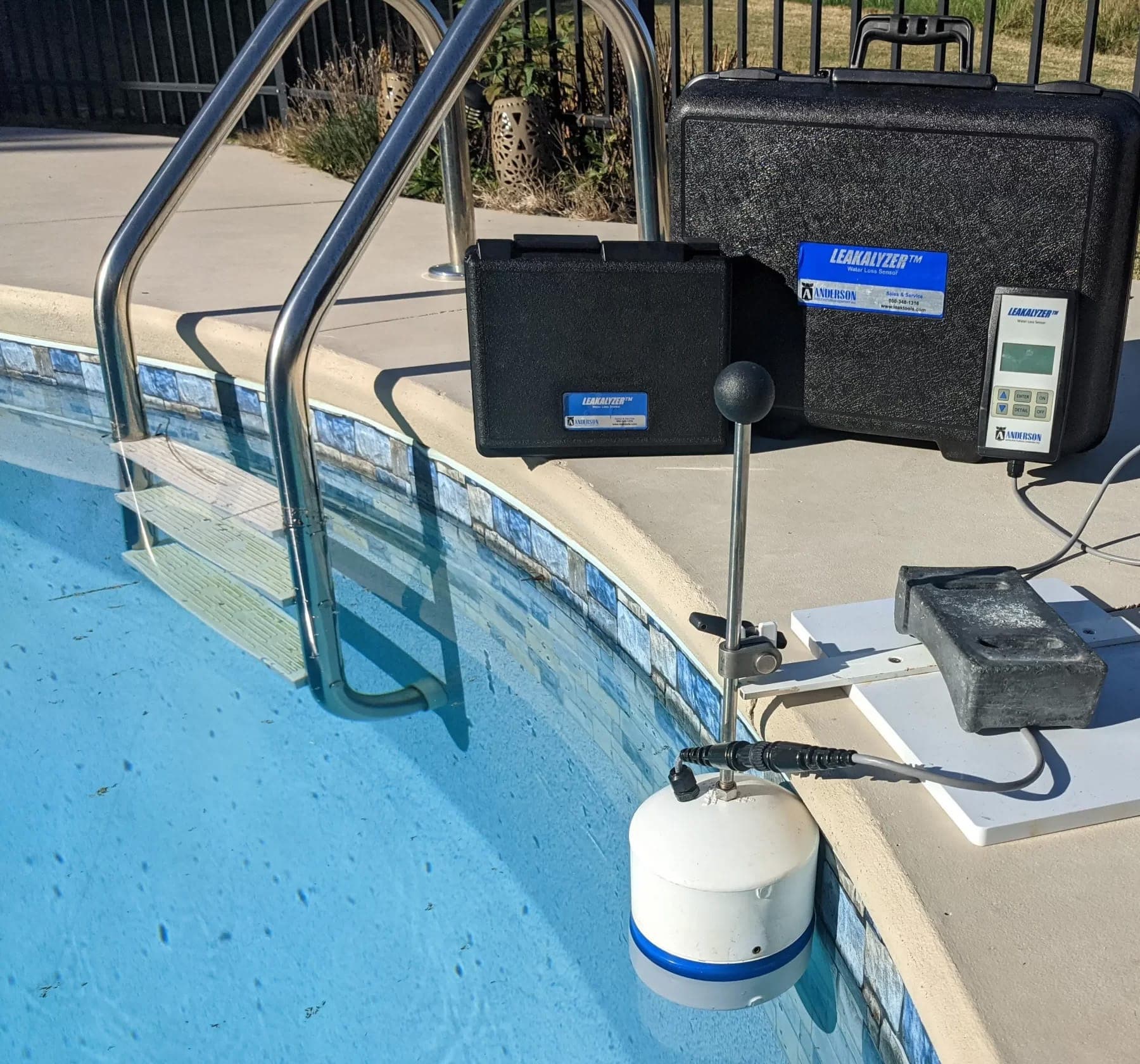 Pool-side water-leak detection rig at the edge of a residential swimming pool in Sierra County, a LEAKALYZER water-loss sensor mounted on a tripod stand, a probe sensor submerged in the pool, equipment cases on the deck, and the pool's tile coping and ladder visible, a precise diagnostic measuring water loss to pinpoint leaks in the pool shell or plumbing.