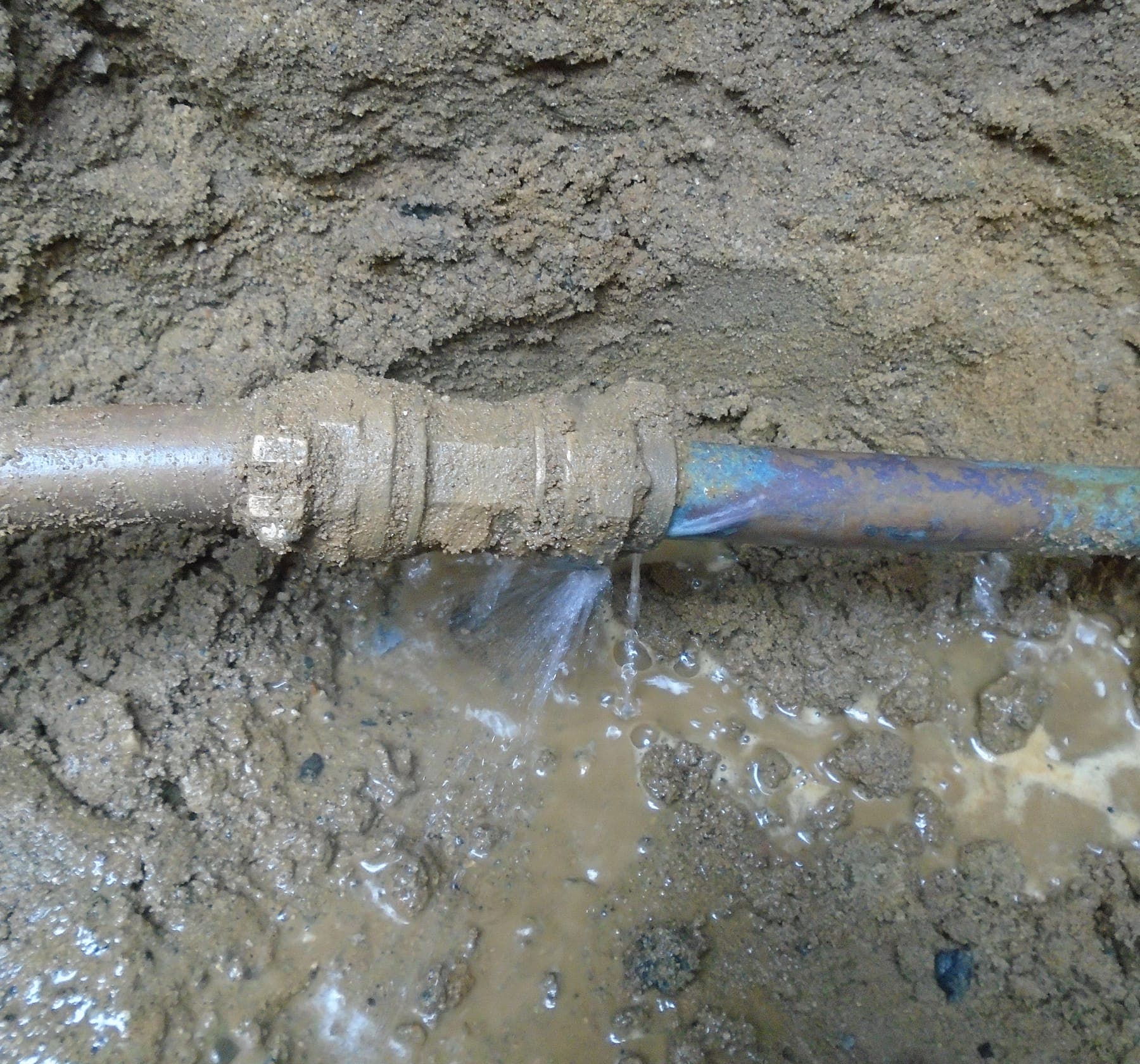 Buried copper water line leaking from a coupling in muddy soil.