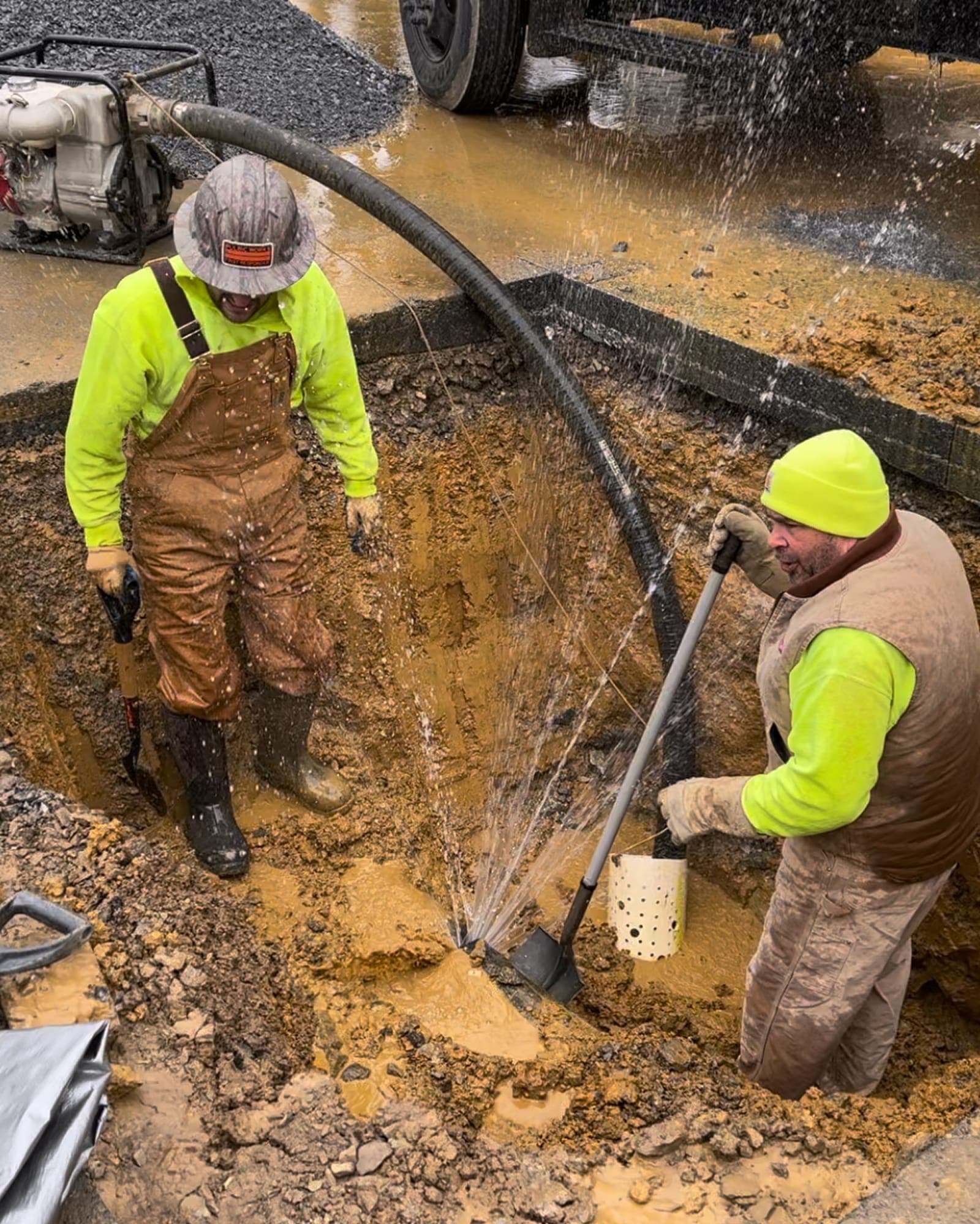 Two workers in a muddy excavation beside a pressurized underground water line spraying from an active leak.