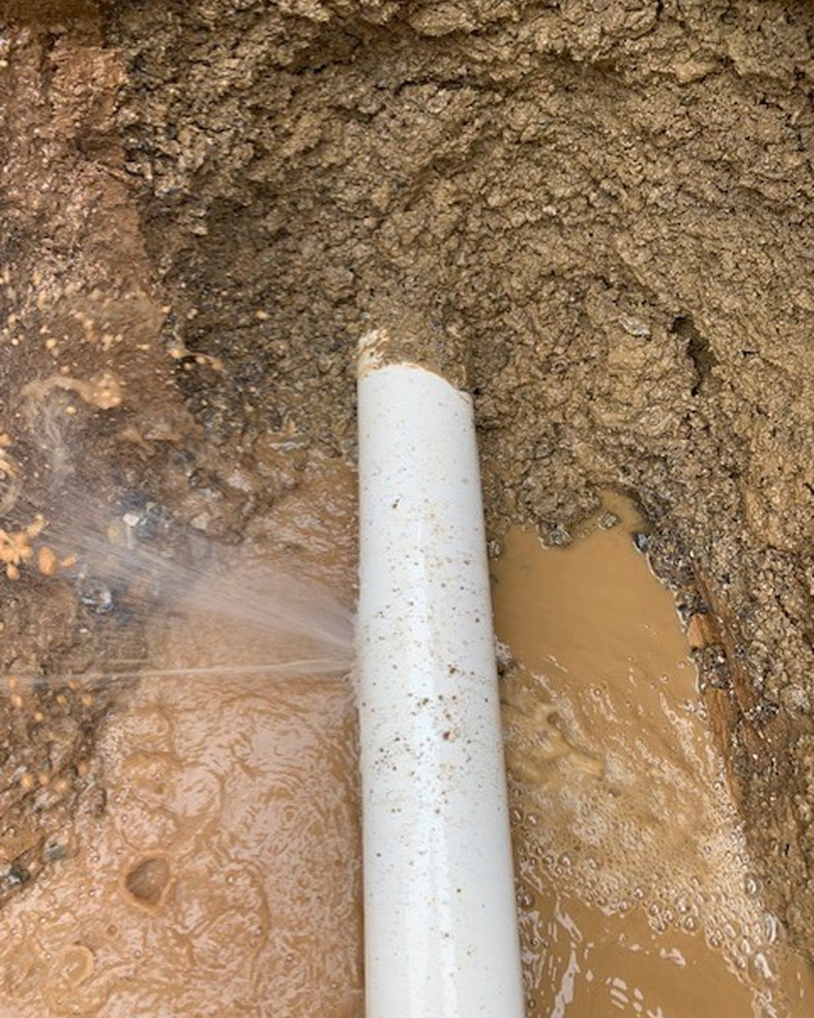 White sewer pipe leaking inside a muddy trench.